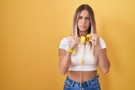 Téléchargez les photos : Jeune femme blonde debout sur fond jaune portant des écouteurs prêts à se battre avec geste de défense poing, visage en colère et contrarié, peur du problème - en image libre de droit