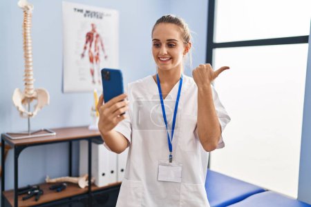 Photo for Young physiotherapist woman working at pain recovery clinic doing video appointment pointing thumb up to the side smiling happy with open mouth - Royalty Free Image