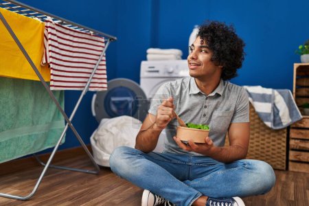 Photo for Young hispanic man eating salad waiting for washing machine at laundry room - Royalty Free Image
