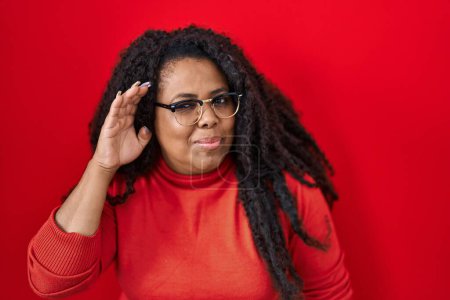 Photo for Plus size hispanic woman standing over red background smiling with hand over ear listening an hearing to rumor or gossip. deafness concept. - Royalty Free Image