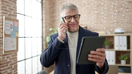 Photo for Middle age grey-haired man business worker talking on smartphone using touchpad at office - Royalty Free Image