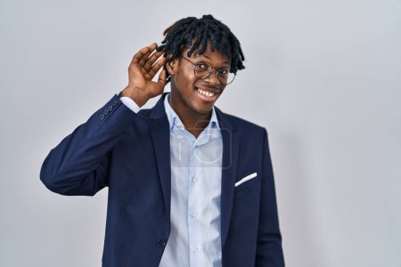 Téléchargez les photos : Jeune homme africain avec dreadlocks veste d'affaires sur fond blanc souriant avec la main sur l'oreille en écoutant une rumeur ou des commérages. concept de surdité. - en image libre de droit