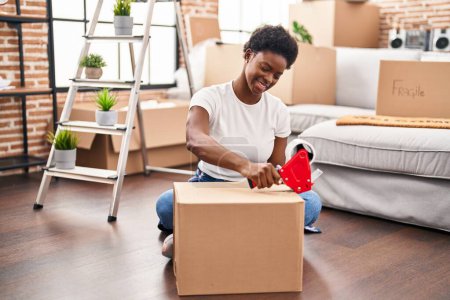 Téléchargez les photos : Femme afro-américaine emballage boîte en carton assis sur le sol à la nouvelle maison - en image libre de droit