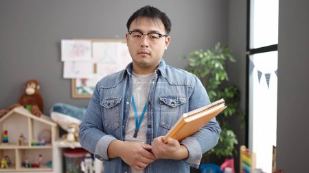 Photo for Young chinese man teacher holding books with relaxed expression at kindergarten - Royalty Free Image