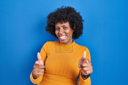 Téléchargez les photos : Femme noire avec des cheveux bouclés debout sur fond bleu pointant les doigts vers la caméra avec un visage heureux et drôle. bonne énergie et vibes. - en image libre de droit