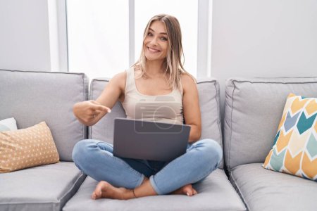 Téléchargez les photos : Blonde femme caucasienne en utilisant un ordinateur portable à la maison assis sur le canapé souriant heureux pointant avec la main et le doigt - en image libre de droit