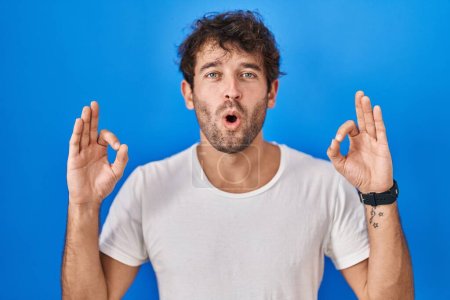 Photo for Hispanic young man standing over blue background looking surprised and shocked doing ok approval symbol with fingers. crazy expression - Royalty Free Image