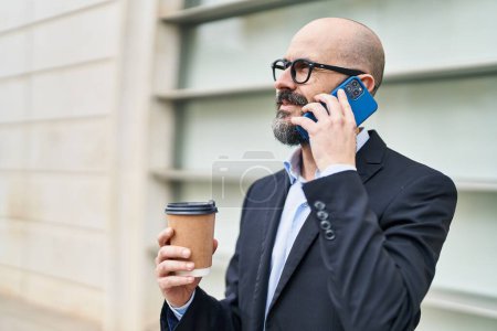 Foto de Joven hombre calvo trabajador de negocios hablando en teléfono inteligente beber café en la calle - Imagen libre de derechos