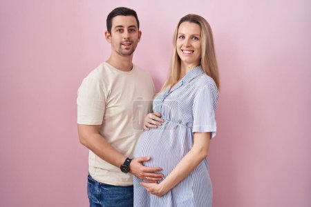 Téléchargez les photos : Jeune couple s'attendant à un bébé debout sur fond rose avec un sourire heureux et cool sur le visage. chanceux. - en image libre de droit