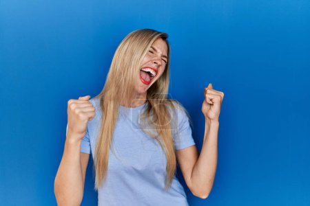 Foto de Hermosa mujer rubia vistiendo camiseta casual sobre fondo azul muy feliz y emocionada haciendo gesto ganador con los brazos levantados, sonriendo y gritando por el éxito. concepto de celebración. - Imagen libre de derechos