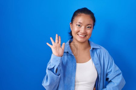 Téléchargez les photos : Asiatique jeune femme debout sur fond bleu renonçant à dire bonjour heureux et souriant, geste de bienvenue amical - en image libre de droit