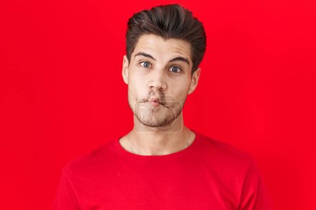 Photo for Young hispanic man standing over red background making fish face with lips, crazy and comical gesture. funny expression. - Royalty Free Image