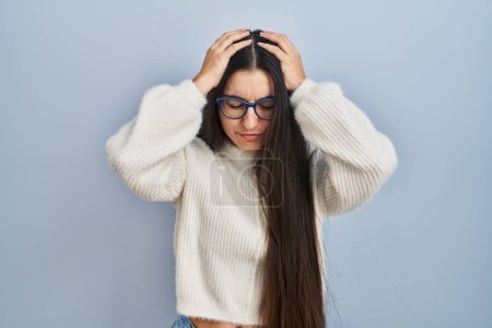 Foto de Mujer hispana joven con suéter casual sobre fondo azul que sufre de dolor de cabeza desesperada y estresada debido al dolor y la migraña. manos en la cabeza. - Imagen libre de derechos