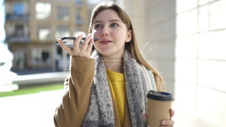 Foto de Young blonde woman talking on smartphone drinking coffee at street - Imagen libre de derechos