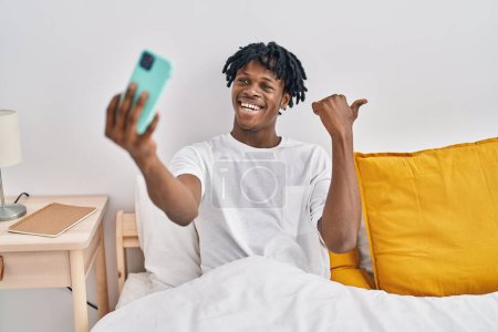Téléchargez les photos : Jeune homme africain avec dreadlocks prendre un selfie sur le lit pointant le pouce vers le côté souriant heureux avec la bouche ouverte - en image libre de droit