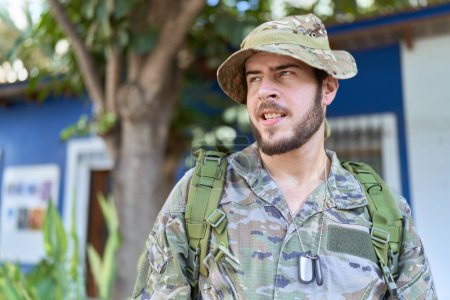 Téléchargez les photos : Jeune homme hispanique portant l'uniforme de camouflage de l'armée à l'extérieur regardant de côté avec le sourire sur le visage, expression naturelle. riant confiant. - en image libre de droit