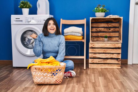 Foto de Joven mujer hispana en la lavandería apuntando con el pulgar hacia el lado sonriendo feliz con la boca abierta - Imagen libre de derechos