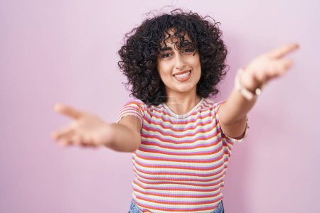Téléchargez les photos : Jeune femme du Moyen-Orient debout sur fond rose souriant joyeuse offrant des mains donnant assistance et acceptation. - en image libre de droit