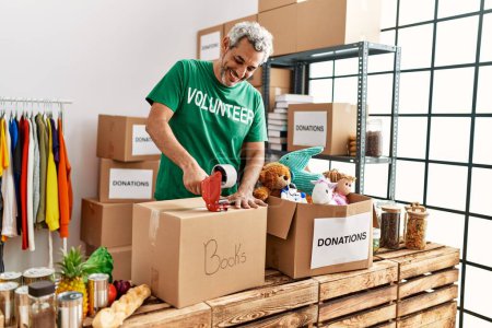Photo for Middle age grey-haired man volunteer smiling confident packing books cardboard box at charity center - Royalty Free Image