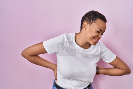 Foto de Hermosa mujer afroamericana de pie sobre fondo rosa sufriendo de dolor de espalda, tocando la espalda con la mano, dolor muscular - Imagen libre de derechos