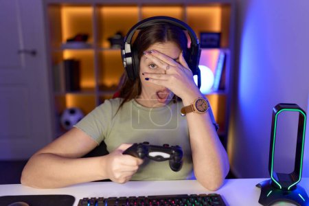 Photo for Beautiful brunette woman playing video games wearing headphones peeking in shock covering face and eyes with hand, looking through fingers with embarrassed expression. - Royalty Free Image