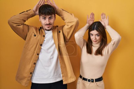 Photo for Young hispanic couple standing over yellow background doing bunny ears gesture with hands palms looking cynical and skeptical. easter rabbit concept. - Royalty Free Image