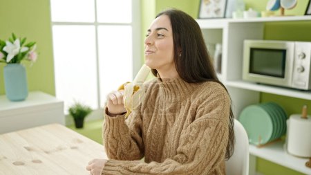 Foto de Joven hermosa mujer hispana comiendo plátano sentado en la mesa en la cocina - Imagen libre de derechos