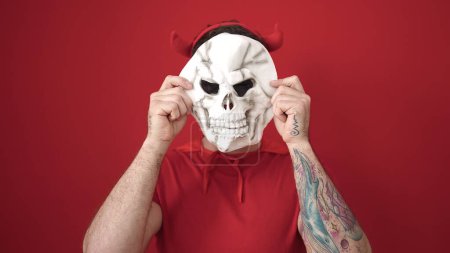 Téléchargez les photos : Jeune homme caucasien portant costume diable couvrant visage avec crâne sur fond rouge isolé - en image libre de droit