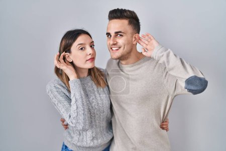 Téléchargez les photos : Jeune couple hispanique debout sur fond blanc souriant avec la main sur l'oreille en écoutant une rumeur ou des commérages. concept de surdité. - en image libre de droit