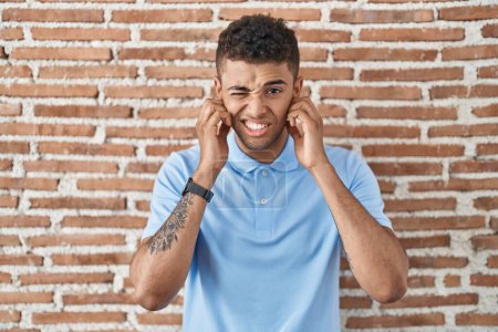 Téléchargez les photos : Jeune homme brésilien debout sur un mur de briques couvrant les oreilles avec des doigts avec une expression agacée pour le bruit de la musique forte. concept sourd. - en image libre de droit