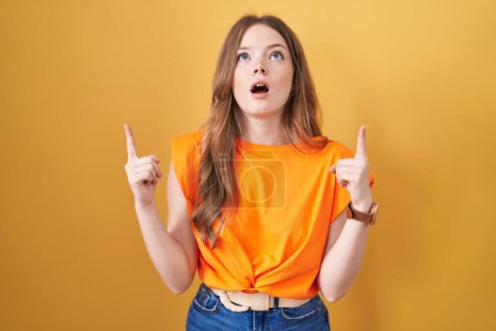 Foto de Caucasian woman standing over yellow background amazed and surprised looking up and pointing with fingers and raised arms. - Imagen libre de derechos