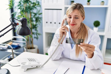 Foto de Young blonde woman wearing dentist uniform talking on the telephone holding xray at clinic - Imagen libre de derechos
