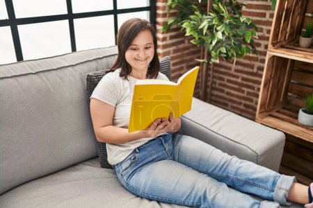 Foto de Síndrome de Down mujer leyendo libro sentado en el sofá en casa - Imagen libre de derechos
