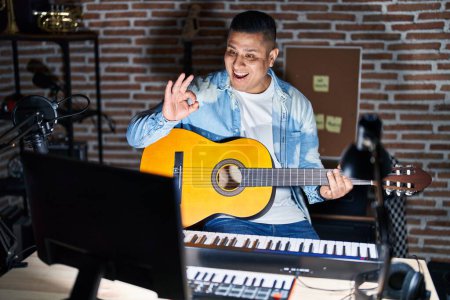 Téléchargez les photos : Jeune homme hispanique jouant de la guitare classique au studio de musique souriant positif faire ok signe avec la main et les doigts. expression réussie. - en image libre de droit