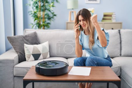 Foto de Joven mujer hispana sentada en casa por robot de vacío hablando por teléfono molesta y frustrada gritando de rabia, gritando loca de rabia y la mano levantada - Imagen libre de derechos