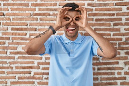 Photo for Brazilian young man standing over brick wall doing ok gesture like binoculars sticking tongue out, eyes looking through fingers. crazy expression. - Royalty Free Image