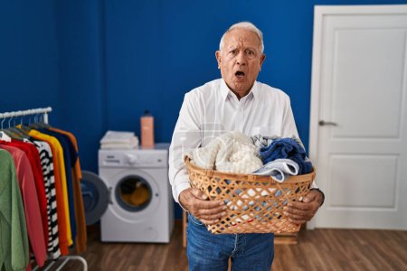 Photo for Senior man with grey hair holding laundry basket at home in shock face, looking skeptical and sarcastic, surprised with open mouth - Royalty Free Image