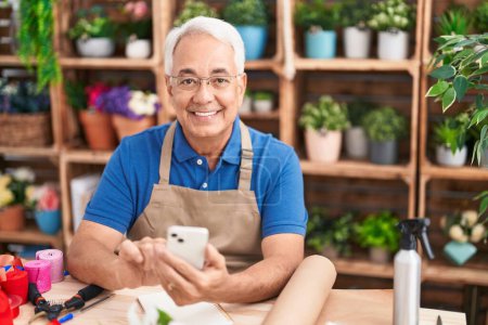 Foto de Florista hombre de pelo gris de mediana edad sonriendo confiado usando teléfono inteligente en floristería - Imagen libre de derechos