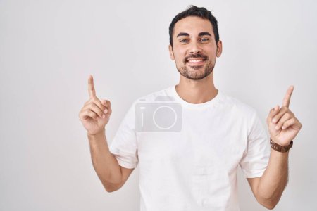 Foto de Hombre hispano guapo de pie sobre fondo blanco sonriendo asombrado y sorprendido y señalando con los dedos y los brazos levantados. - Imagen libre de derechos