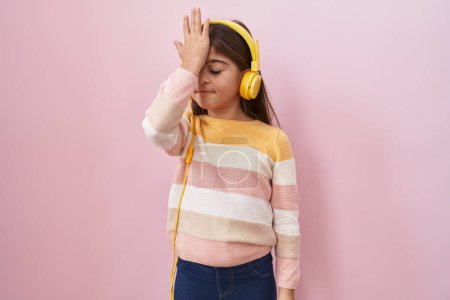 Téléchargez les photos : Petite fille hispanique écoutant de la musique à l'aide d'écouteurs surpris avec la main sur la tête pour erreur, rappelez-vous erreur. oublié, mauvais concept de mémoire. - en image libre de droit
