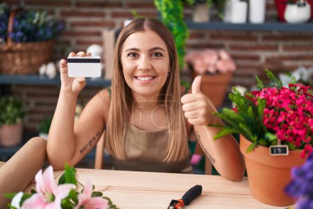 Foto de Mujer rubia joven que trabaja en floristería sosteniendo tarjeta de crédito sonriendo feliz y positivo, pulgar hacia arriba haciendo excelente y signo de aprobación - Imagen libre de derechos