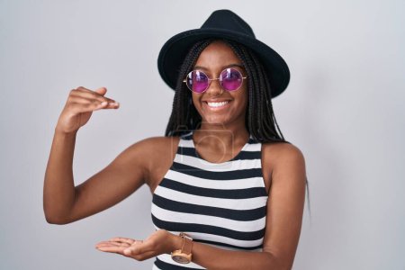 Téléchargez les photos : Jeune afro-américain avec des tresses portant un chapeau et des lunettes de soleil geste avec les mains montrant grand et grand signe de taille, symbole de mesure. souriant en regardant la caméra. concept de mesure. - en image libre de droit