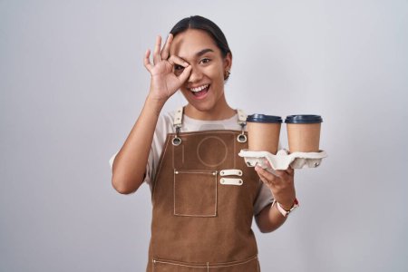 Foto de Mujer hispana joven vistiendo delantal de camarera profesional sosteniendo café haciendo buen gesto con la mano sonriente, ojos mirando a través de los dedos con cara feliz. - Imagen libre de derechos