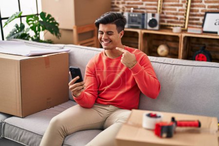 Foto de Hombre hispano mudándose a un nuevo hogar haciendo videollamadas con smartphone sonriendo feliz señalando con la mano y el dedo - Imagen libre de derechos