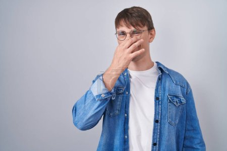 Téléchargez les photos : Homme blond caucasien debout portant des lunettes sentant quelque chose de puant et dégoûtant, odeur intolérable, retenant son souffle avec les doigts sur le nez. mauvaise odeur - en image libre de droit