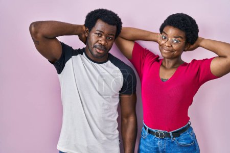 Téléchargez les photos : Jeune couple afro-américain debout sur fond rose relaxant et étirant, bras et mains derrière la tête et le cou souriant heureux - en image libre de droit