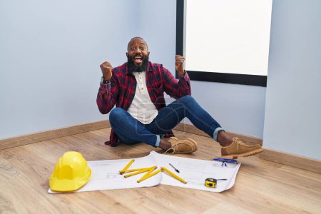 Photo for African american man sitting on the floor at new home looking at blueprints celebrating surprised and amazed for success with arms raised and open eyes. winner concept. - Royalty Free Image