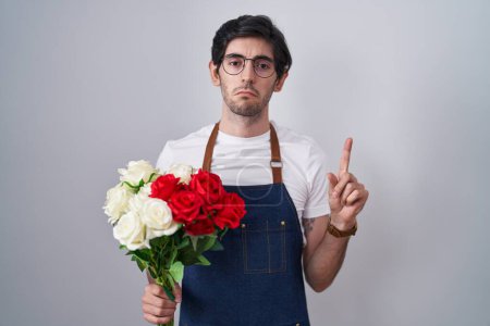 Photo for Young hispanic man holding bouquet of white and red roses pointing up looking sad and upset, indicating direction with fingers, unhappy and depressed. - Royalty Free Image