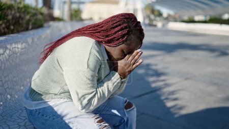 Téléchargez les photos : Femme africaine avec les cheveux tressés regardant triste assis sur un banc au parc - en image libre de droit