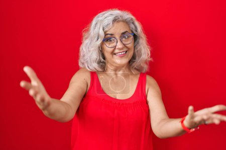 Téléchargez les photos : Femme d'âge moyen avec les cheveux gris debout sur fond rouge souriant joyeuse offrant des mains donnant assistance et acceptation. - en image libre de droit